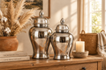 Two silver ginger jars styled casually on a rustic oak shelf with pampas grass and linen books